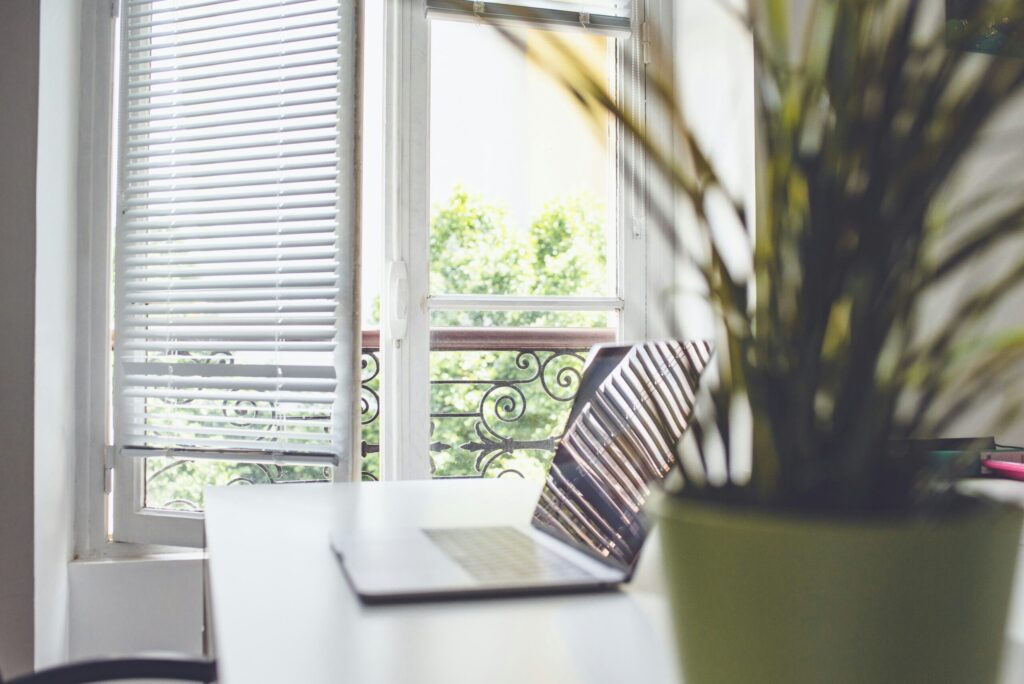 A bright, minimalist office workspace featuring a laptop open on a white desk next to a green potted plant, positioned in front of a large window with white blinds overlooking green trees outside.