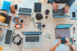Flat lay view of a diverse professional team working around a wooden table with multiple laptops, smartphones, notebooks, and coffee mugs during a collaborative meeting.