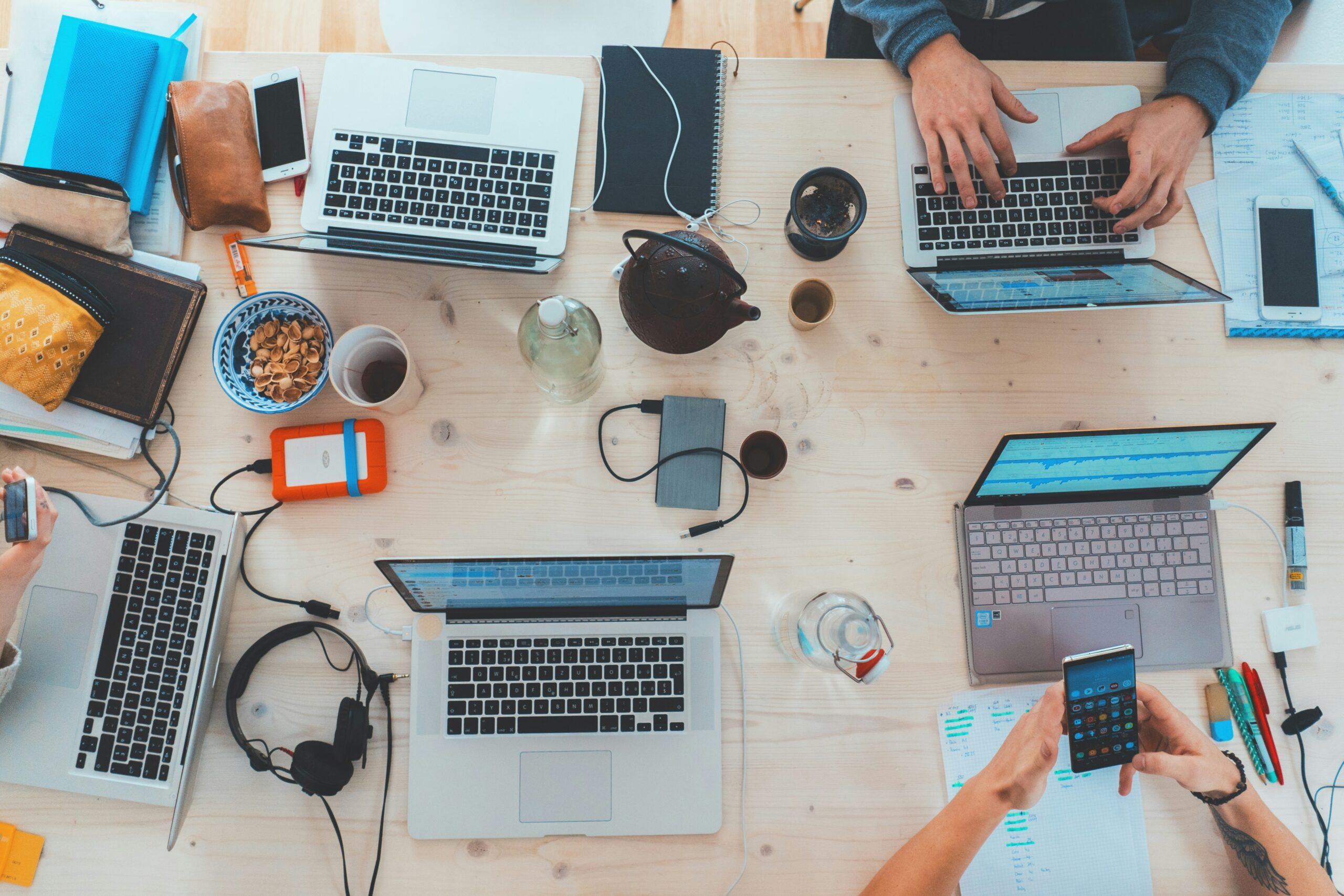 Flat lay view of a diverse professional team working around a wooden table with multiple laptops, smartphones, notebooks, and coffee mugs during a collaborative meeting.